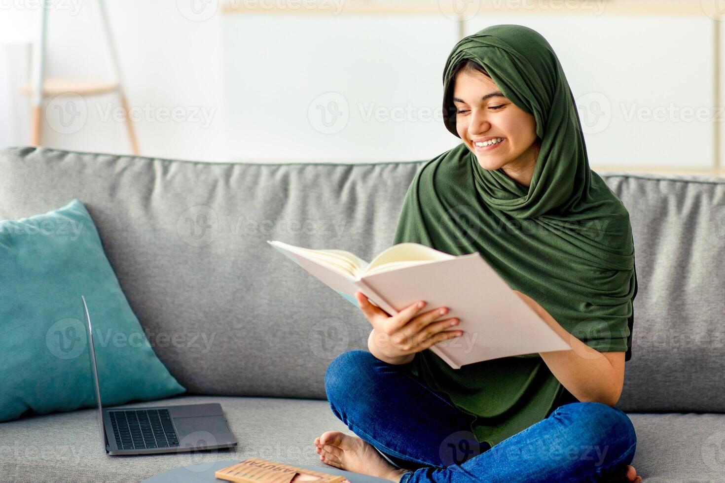 Happy teen girl in hijab sitting on sofa with textbook, reading, studying online from home, preparing for exam. Smart Eastern adolescent in headscarf learning remotely during coronavirus lockdown photo