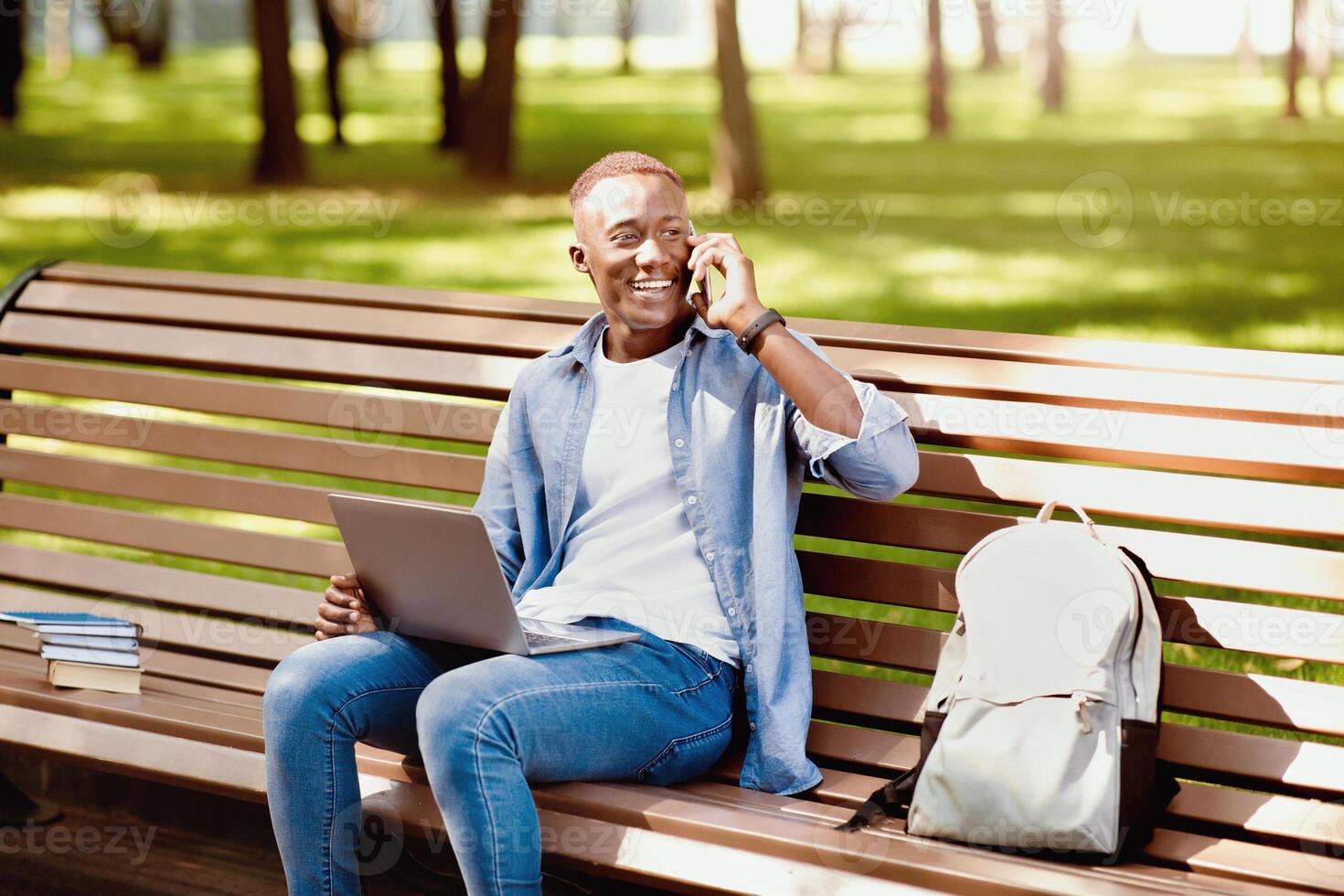 E-learning concept. Funky black student with laptop talking on smartphone on bench at park. African American guy talking to his college tutor while watching educational on pc, outside photo