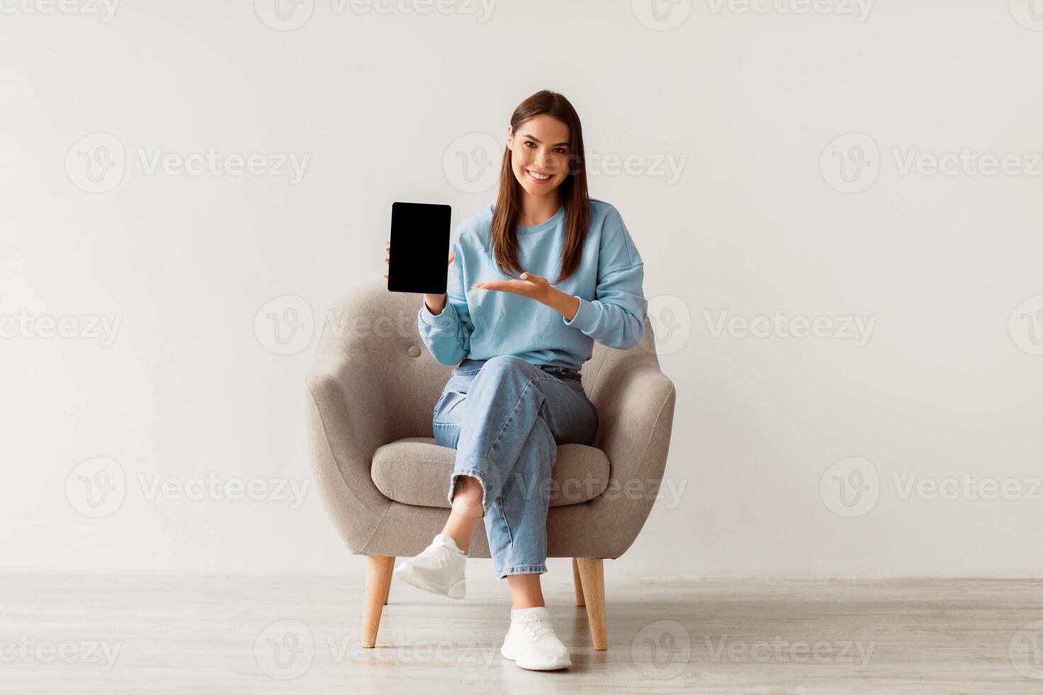 People and gadgets. Happy Caucasian lady showing laptop with blank screen for your website, presenting touch pad with mockup, sitting in armchair against white studio wall, full length photo