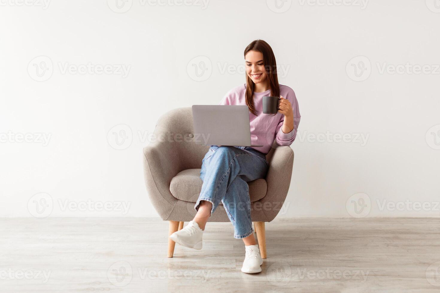 Positive young Caucasian woman working online, sitting in armchair with coffee and using laptop against white studio wall, full length. Cheerful millennial lady surfing internet on portable pc photo