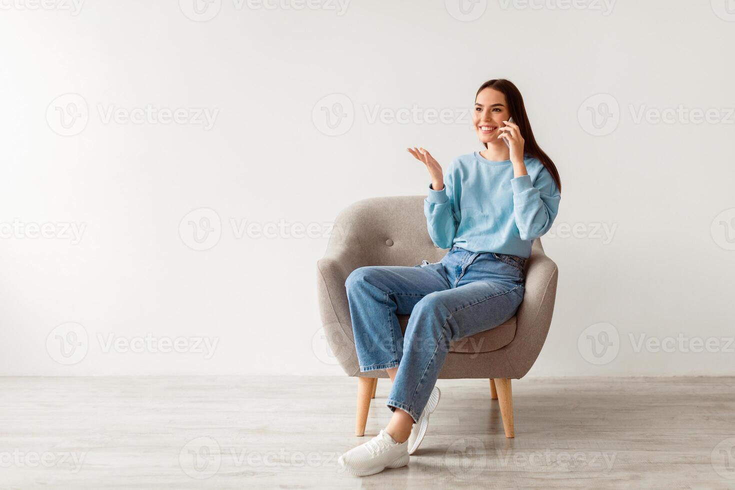 Mobile call. Cheerful young woman talking on cellphone, sitting in armchair against white wall, empty space for ad. Joyful millennial lady having pleasant phone conversation, enjoying communication photo