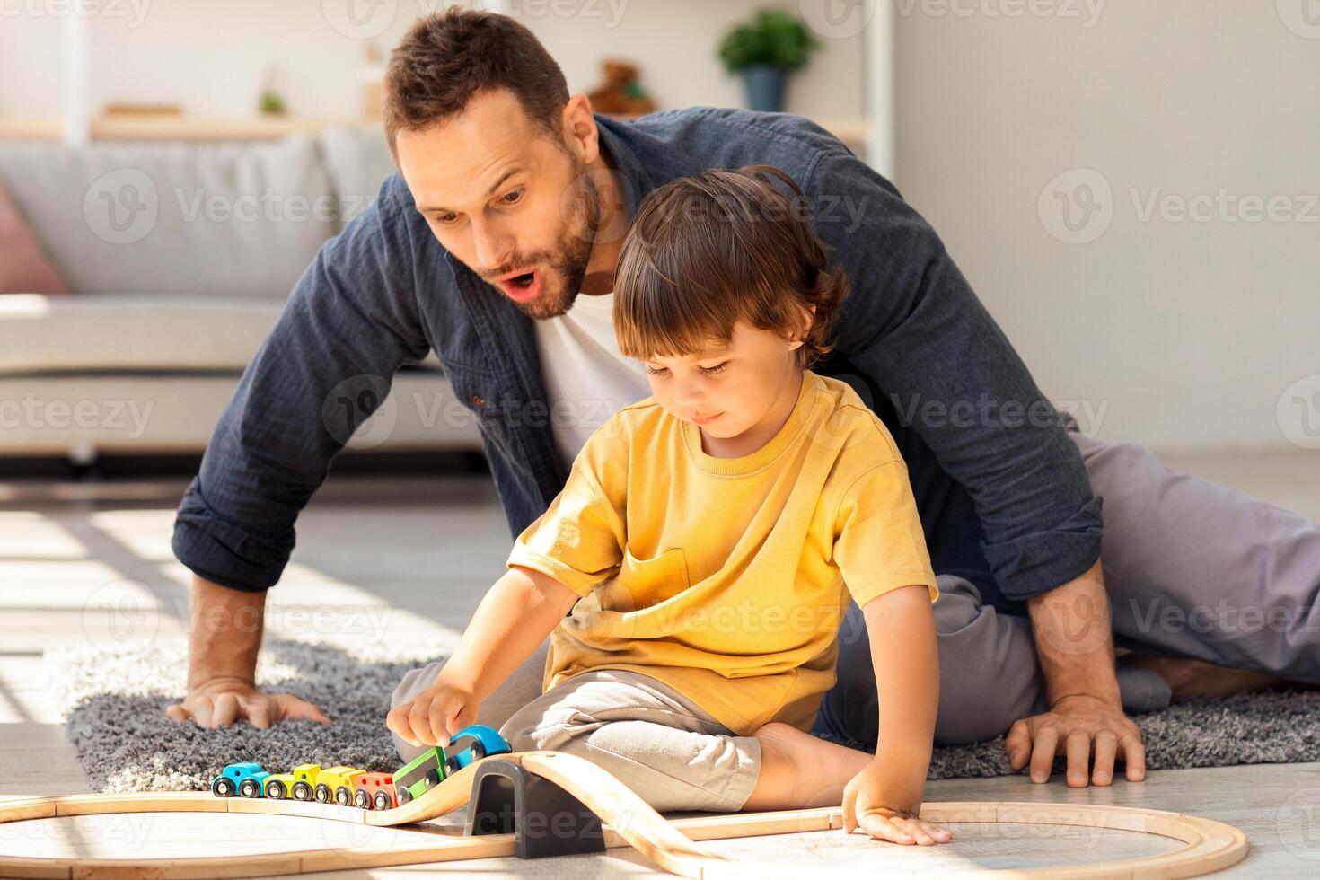 Friendly family concept. Cute little boy playing wooden train with his daddy, surprised man looking at toy with excitement, sitting on floor at home photo