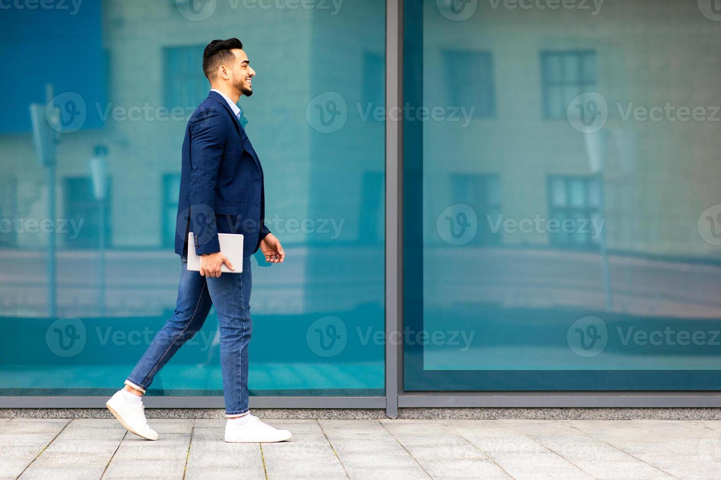 Cheerful young arab guy businessman with pad walking by street, successful middle-eastern entrepreneur or CEO having business meeting, carrying digital tablet, office building background, copy space photo