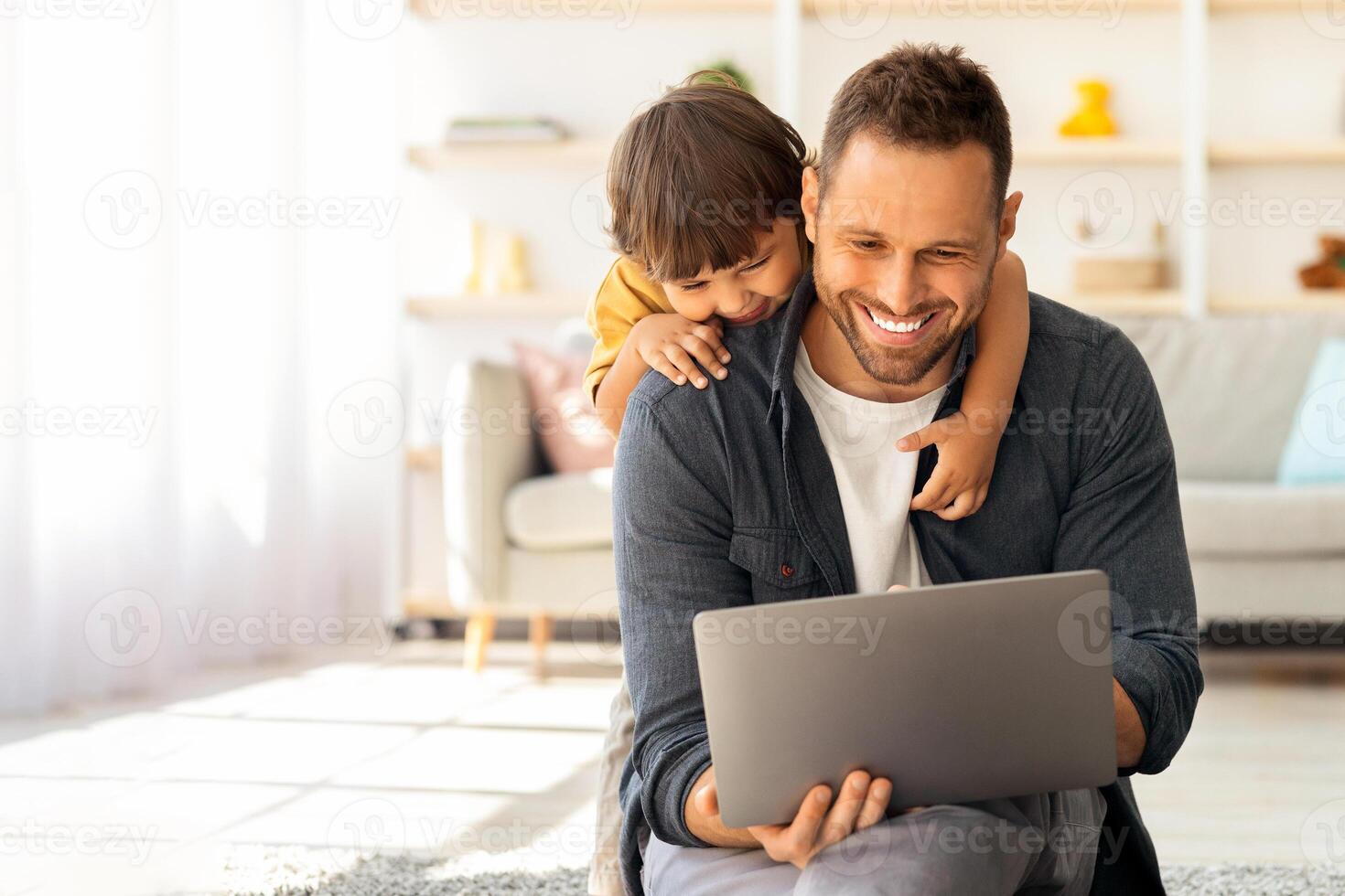 Family business. Smiling man sitting on floor at home and working on laptop, curious little son embracing him and peeking into computer photo