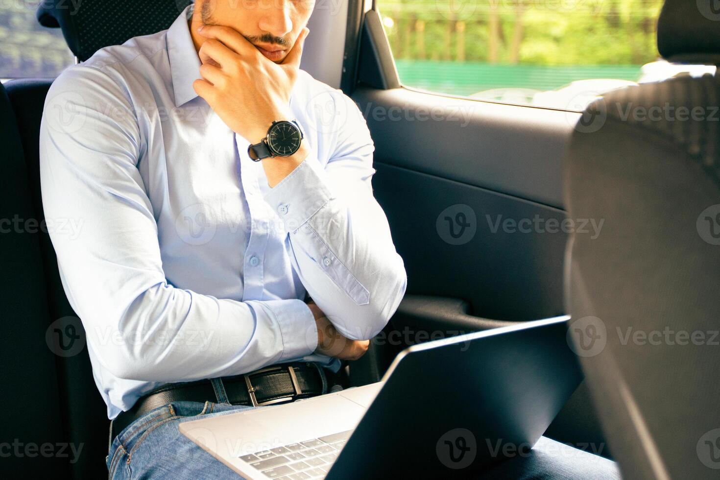 Pensive middle-eastern young businessman working at car back seat, using laptop, looking for creative business solutions while having business trip or going to business meeting, copy space photo