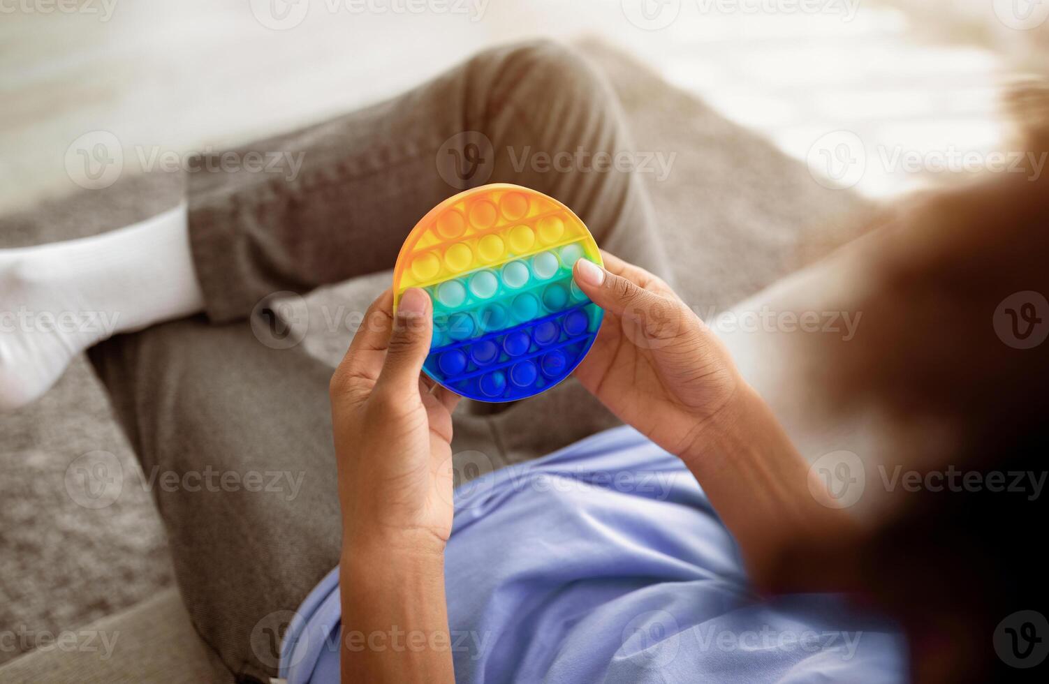Cropped view of black teenager playing with antistress POP IT toy, sitting on sofa at home, closeup. Unrecognizable African American youth enjoying antistress sensory plaything, relaxing indoors photo