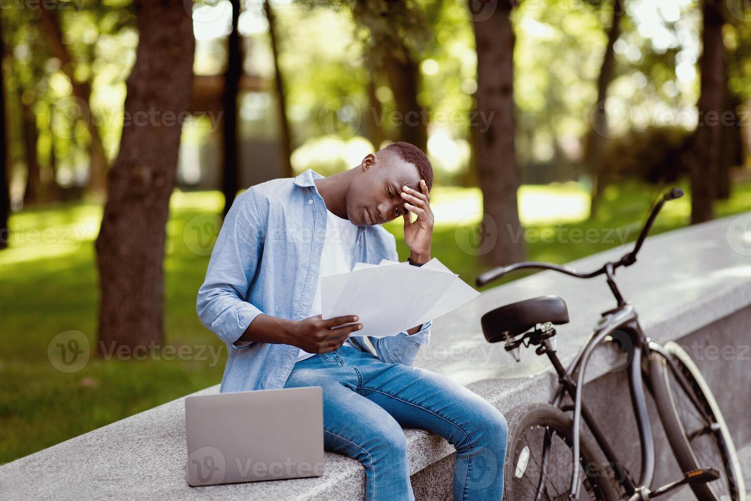 Upset black man with laptop computer and bicycle studying documents at park photo