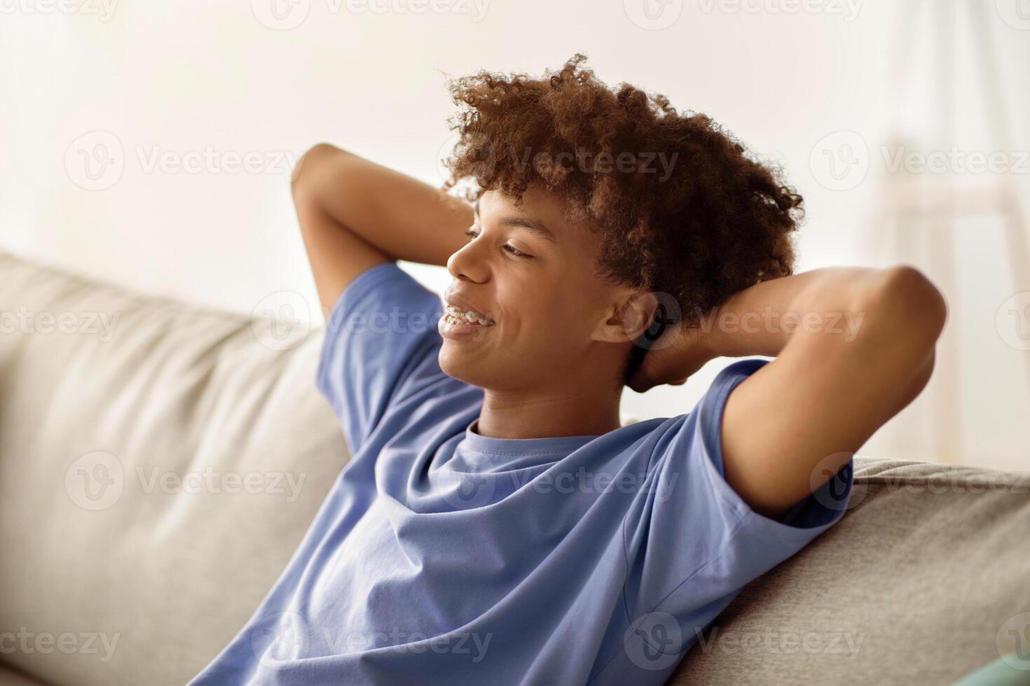 Carefree black teen guy relaxing on couch with hands behind his head, spending time at home during covid lockdown. Peaceful African American youth resting on sofa, having lazy weekend photo