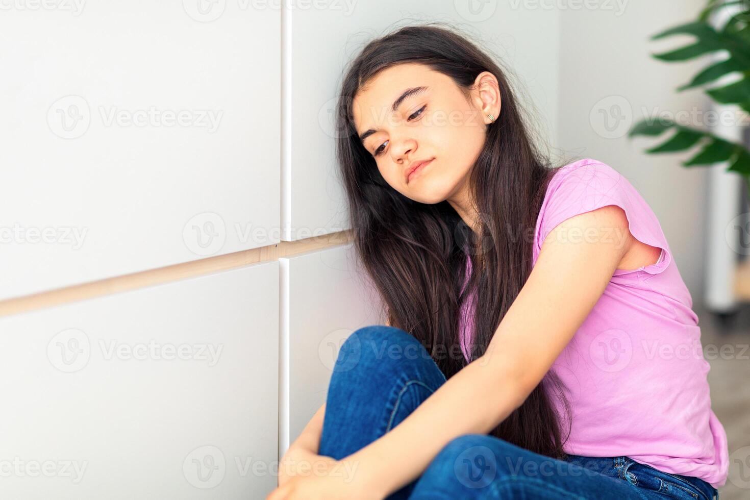 Portrait of Indian teen girl sitting near wall, feeling unhappy and depressed at home. Sad adolescent suffering from loneliness during covid lockdown, missing her friends at times of quarantine photo