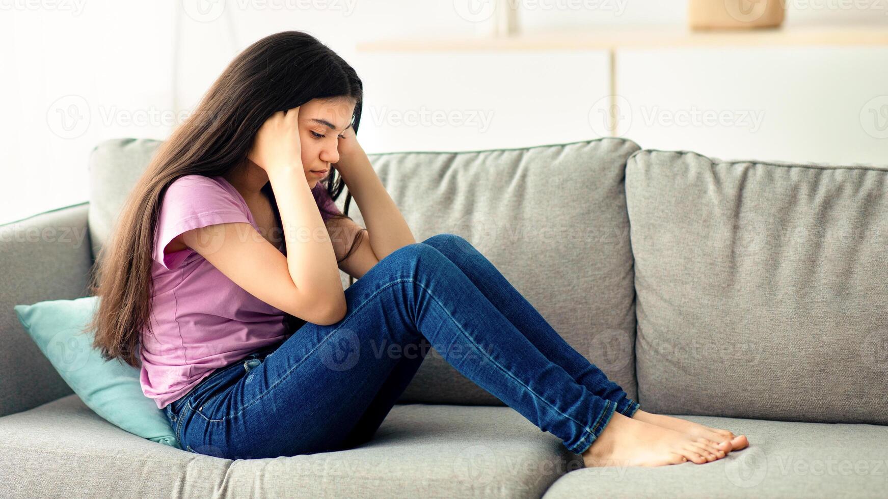 Unhappy teen girl holding her head in despair, feeling depressed, sitting on sofa at home, copy space. Indian asdolescent going through difficult times, having mental problems or mood disorder photo