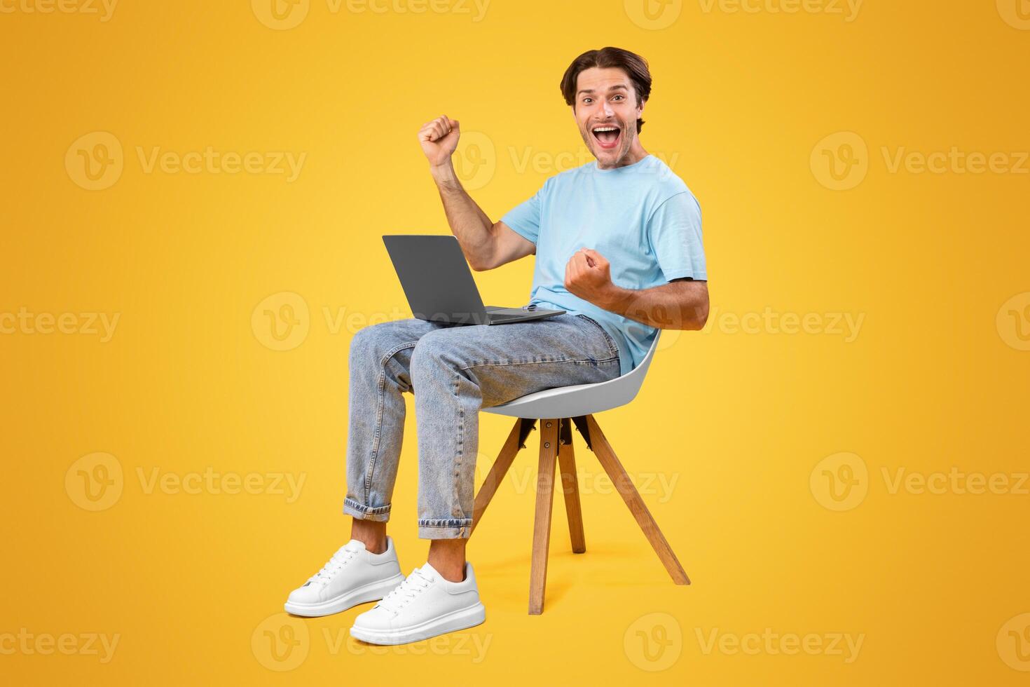 Online Win. Excited Happy Guy Sitting On Chair With Laptop Computer, Emotional Young Man Celebrating Success Shaking Clenched Fists Over Yellow Studio Background, Full Length Shot With Free Space photo