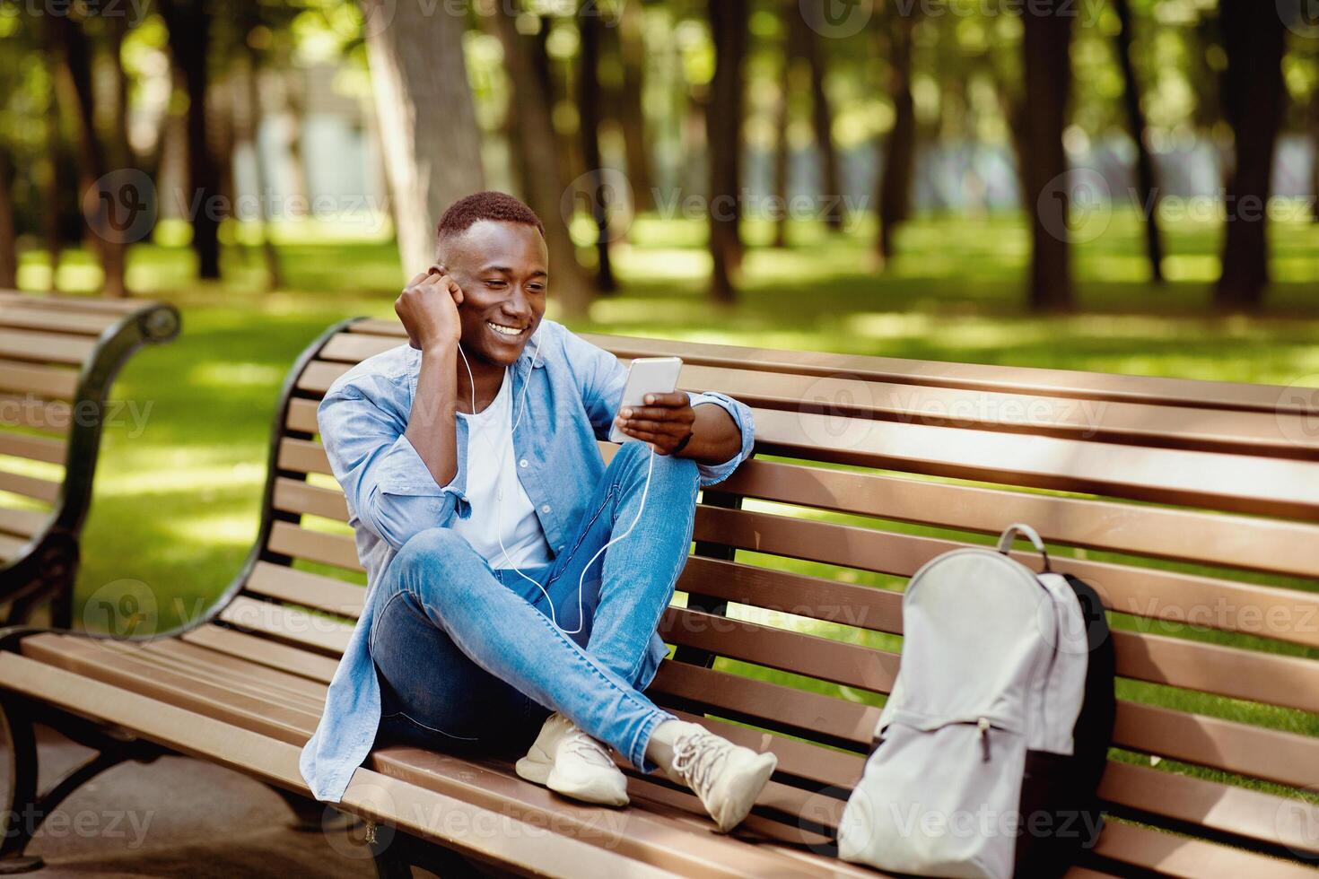 Funky African American guy sitting on bench and enjoying music on mobile phone at urban park, free space photo