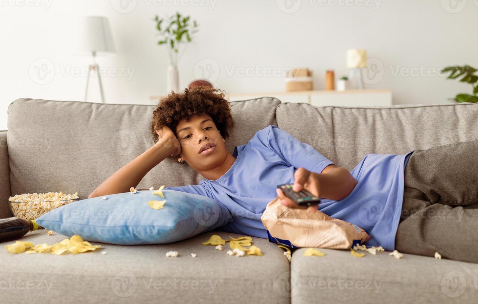 Lazy black teen guy lying on sofa with scattered food, holding TV controller, watching dull movie on television at home. Bored African American adolescent killing time indoors during covid lockdown photo