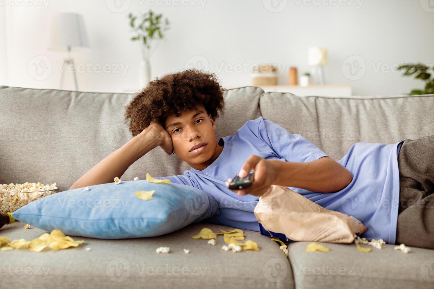 Lazy black teen guy lying on couch with scattered snacks, holding remote control, watching TV at home. Bored African American youth having dull day at home during covid lockdown photo