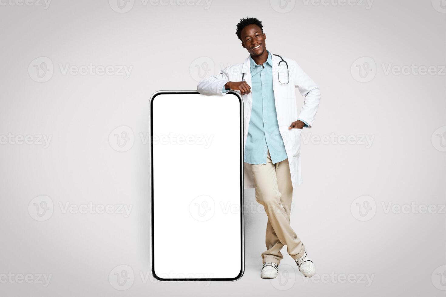 Medical App. African American Male Doctor In Uniform Leaning At Big Blank Smartphone With White Screen, Demonstrating Free Copy Space For Advertisement, Standing On Beige Background, Mockup photo