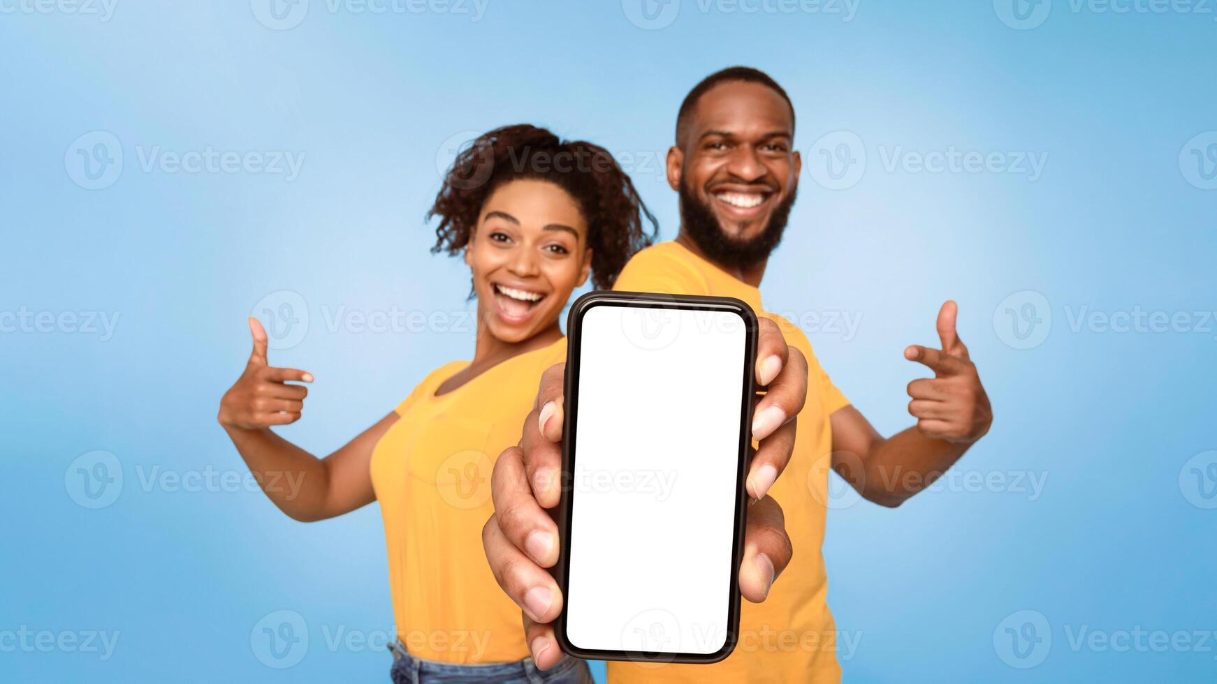 Check this out, cellphone display mockup. Positive African American couple pointing at cellphone with empty screen on blue background, selective focus. Template for mobile app, website or online ad photo
