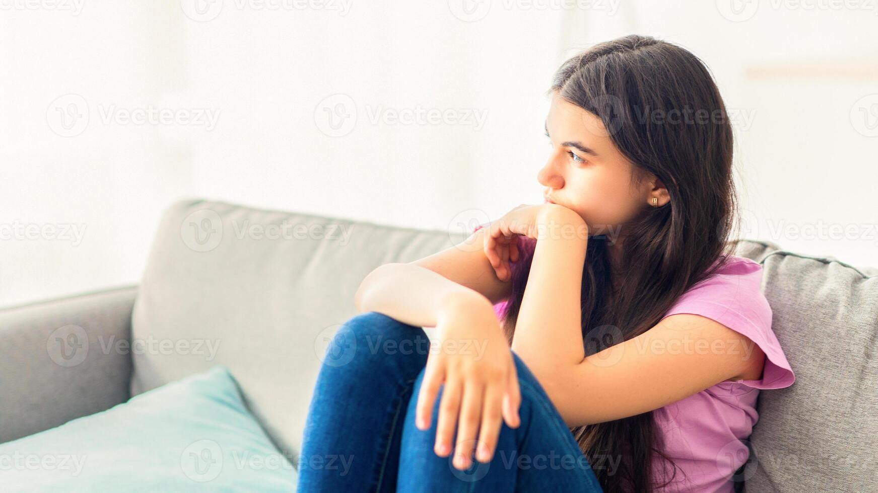 Stressed Indian teenage girl sitting on sofa at home, looking aside, feeling upset and lonely, suffering from lack of communication during covid lockdown. Teen depression concept photo