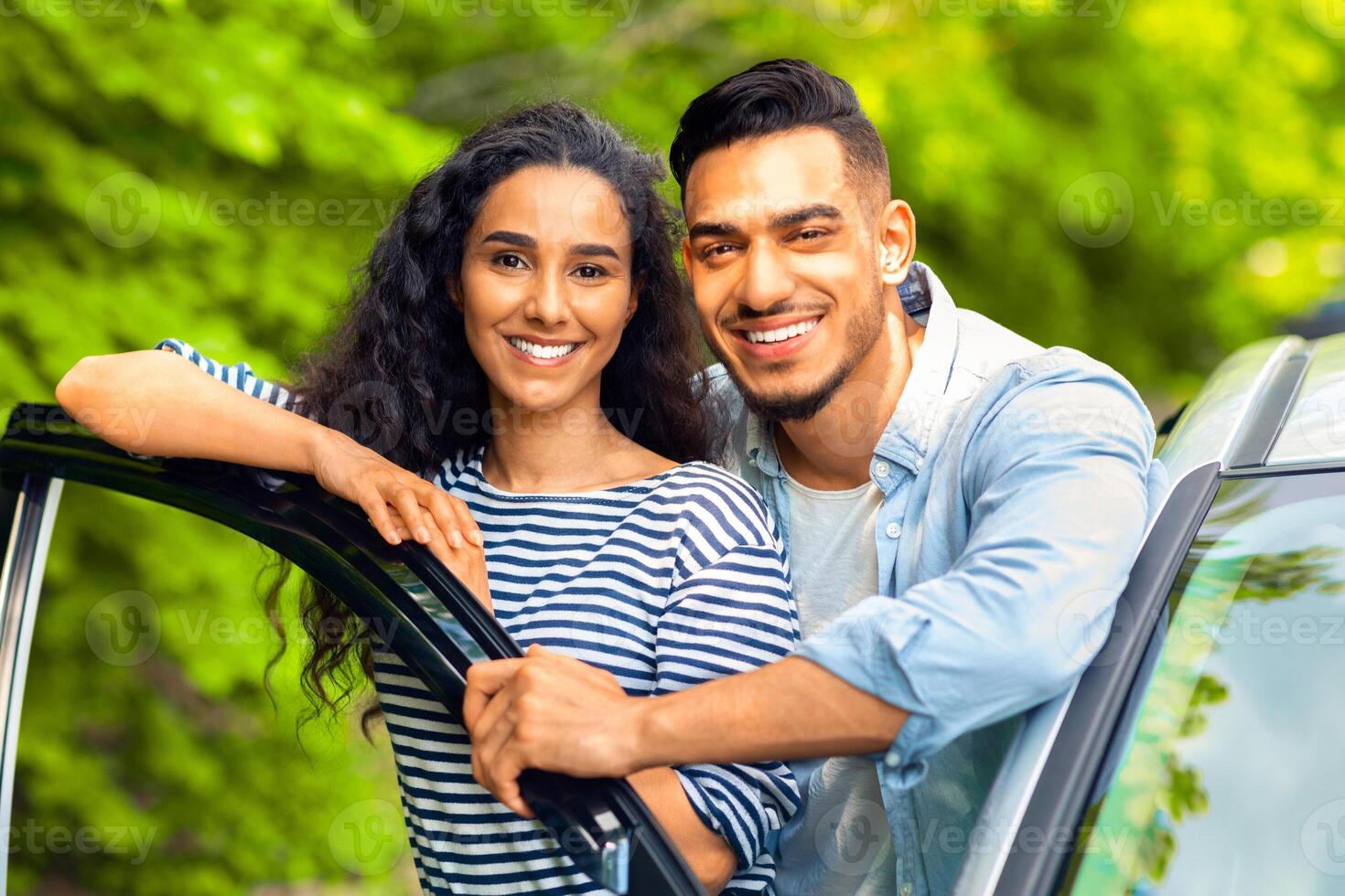 Happy attractive multiracial lovers enjoying car trip to countryside, closeup portrait. Handsome arab guy hugging his pretty woman, standing by open auto door, hugging and smiling at camera photo
