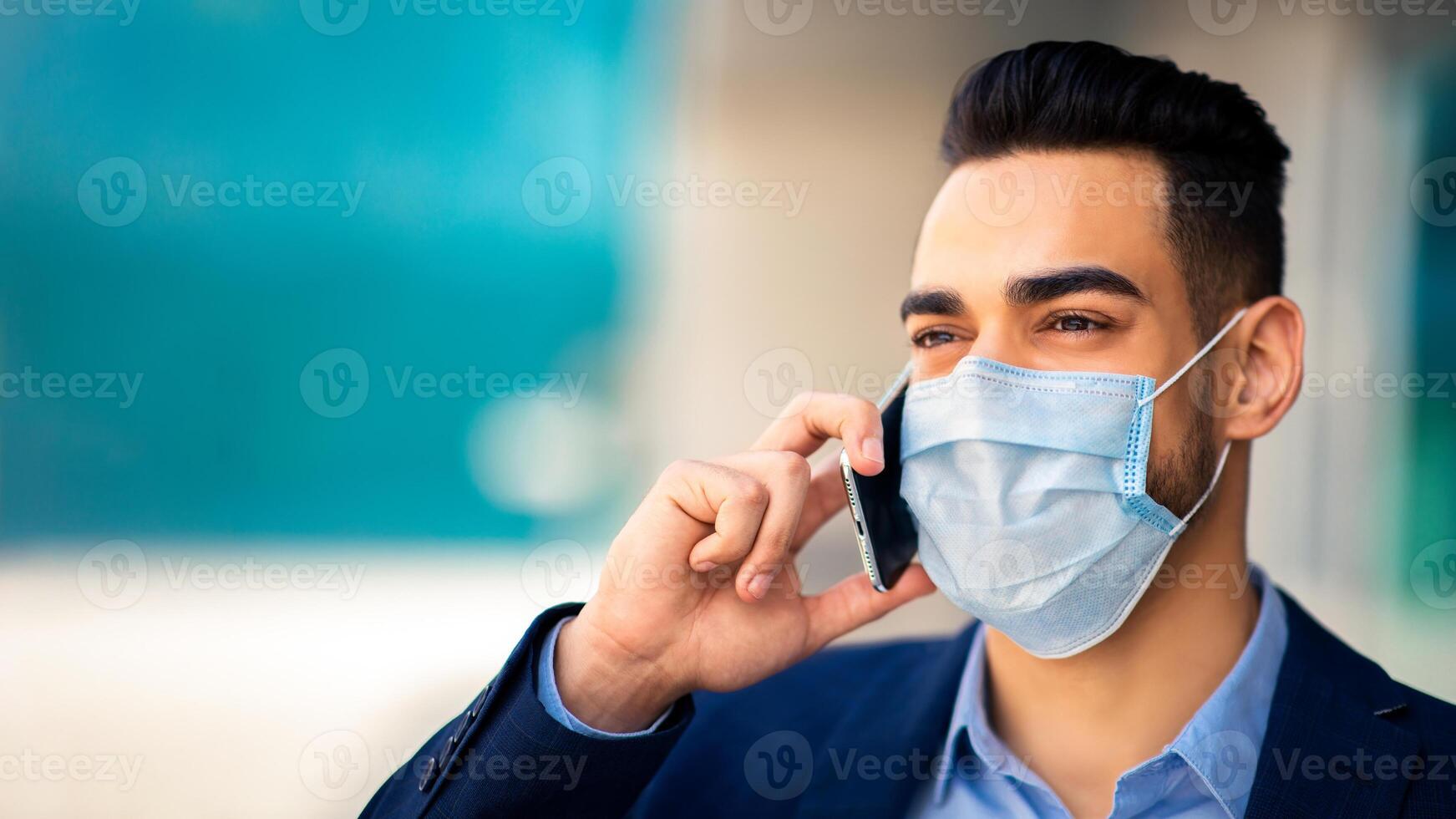 Closeup portrait of young arab businessman in protective medical face mask talking on cellphone next to business center, copy space. Muslim entrepreneur or CEO working during COVID-19 pandemic photo