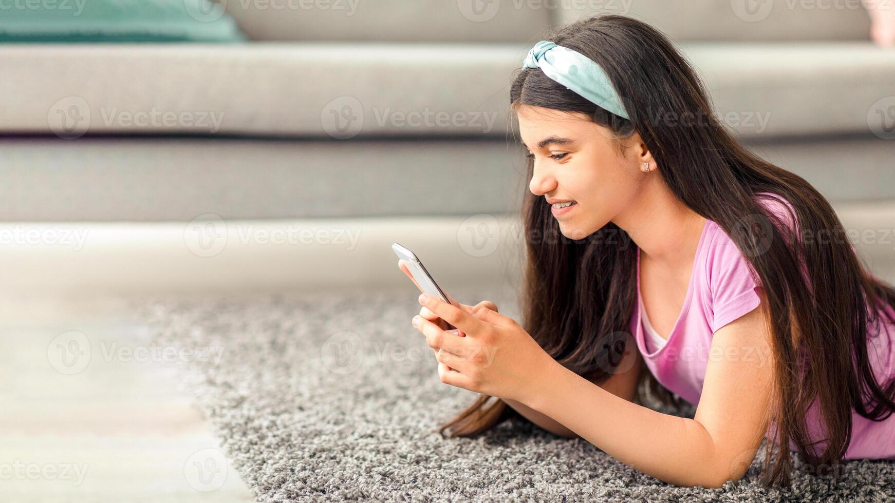 Indian teenage girl with cellphone studying online, chatting with friend, posting in social media at home, free space. Happy teen talking to teacher on web, lying on carpet, using modern gadget photo