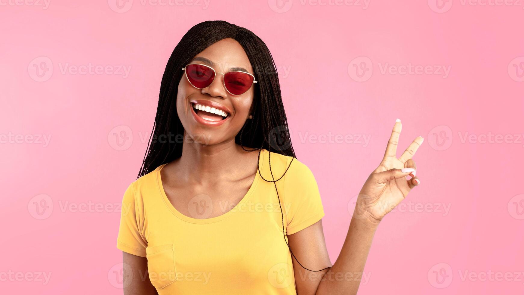 Joyful Black Woman Gesturing Victory Sign To Camera Smiling Posing Over Blue Background, Wearing Sunglasses. Studio Shot Of Happy African American Female Showing Peace Or V-Sign Concept photo