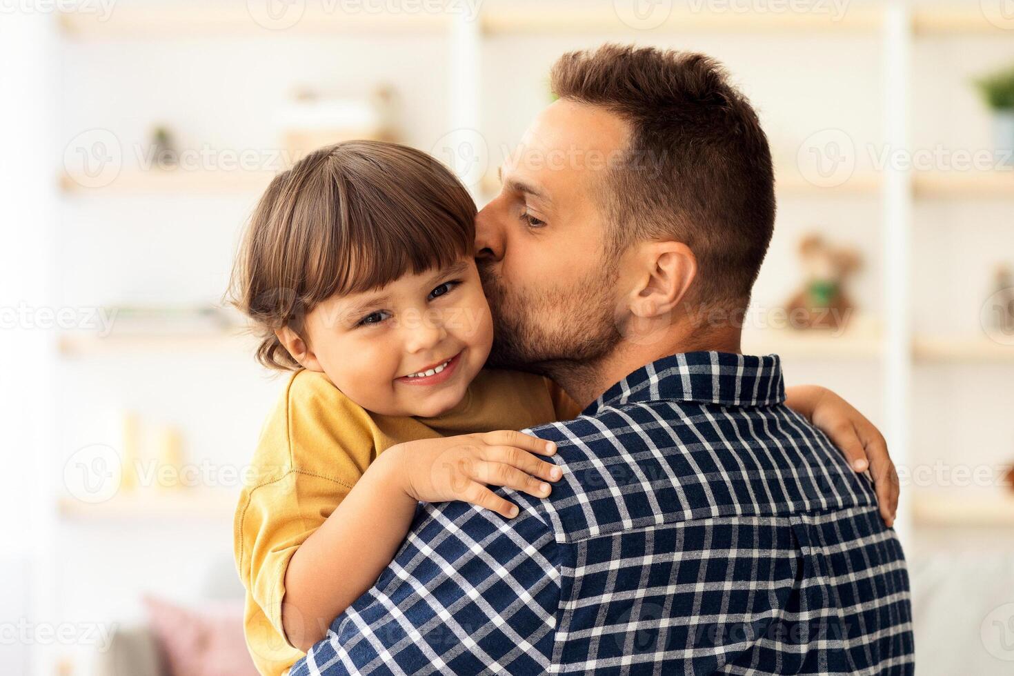 Indoors portrait of happy young father kissing his lovely adorable little son, enjoying time together at home interior, man carrying boy on hands photo