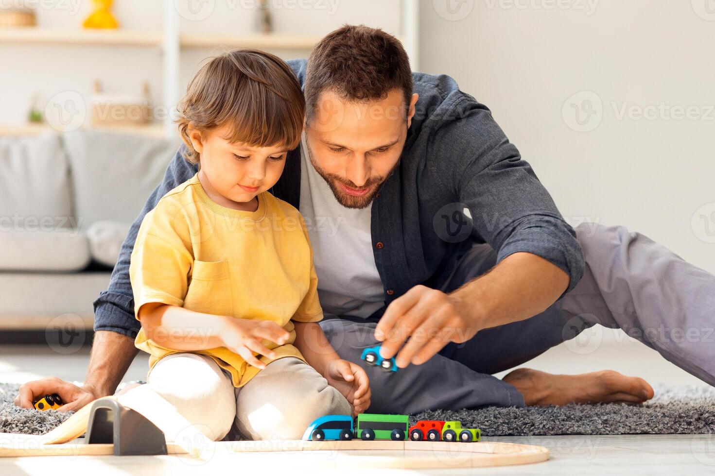 Family fun and leisure. Cute little boy playing wooden railway and trains with his daddy, interested man spending time with kid, sitting on floor at home photo