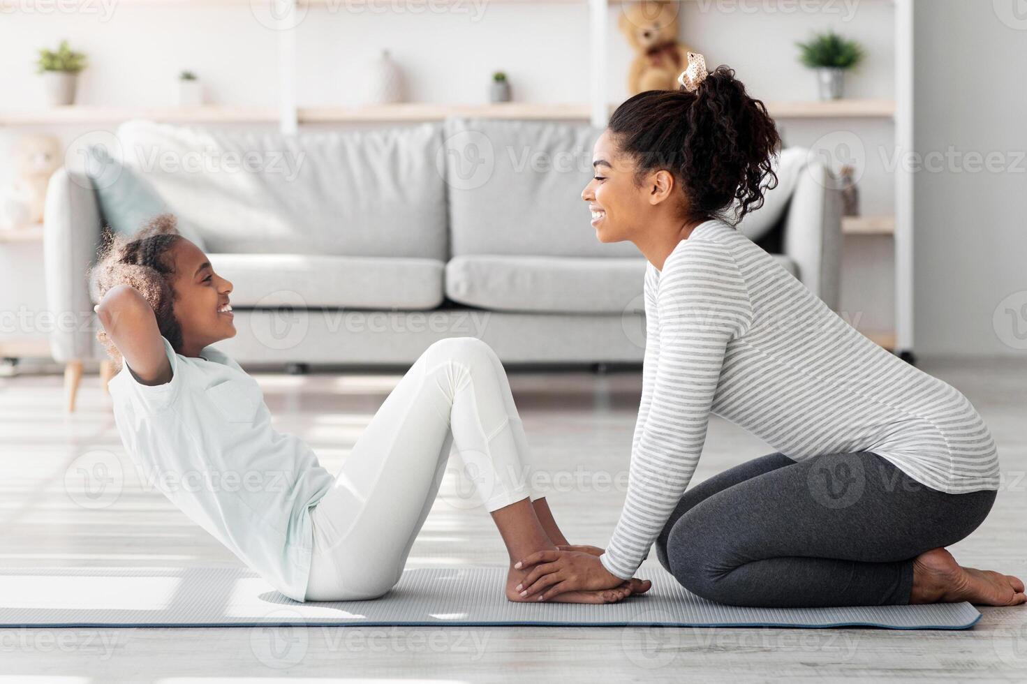 Black mother and daughter in sportswear exercising together at home, sitting on fitness mat and working on abdominal muscles, doing crunches, side view. Sporty family mom and kid doing work out photo