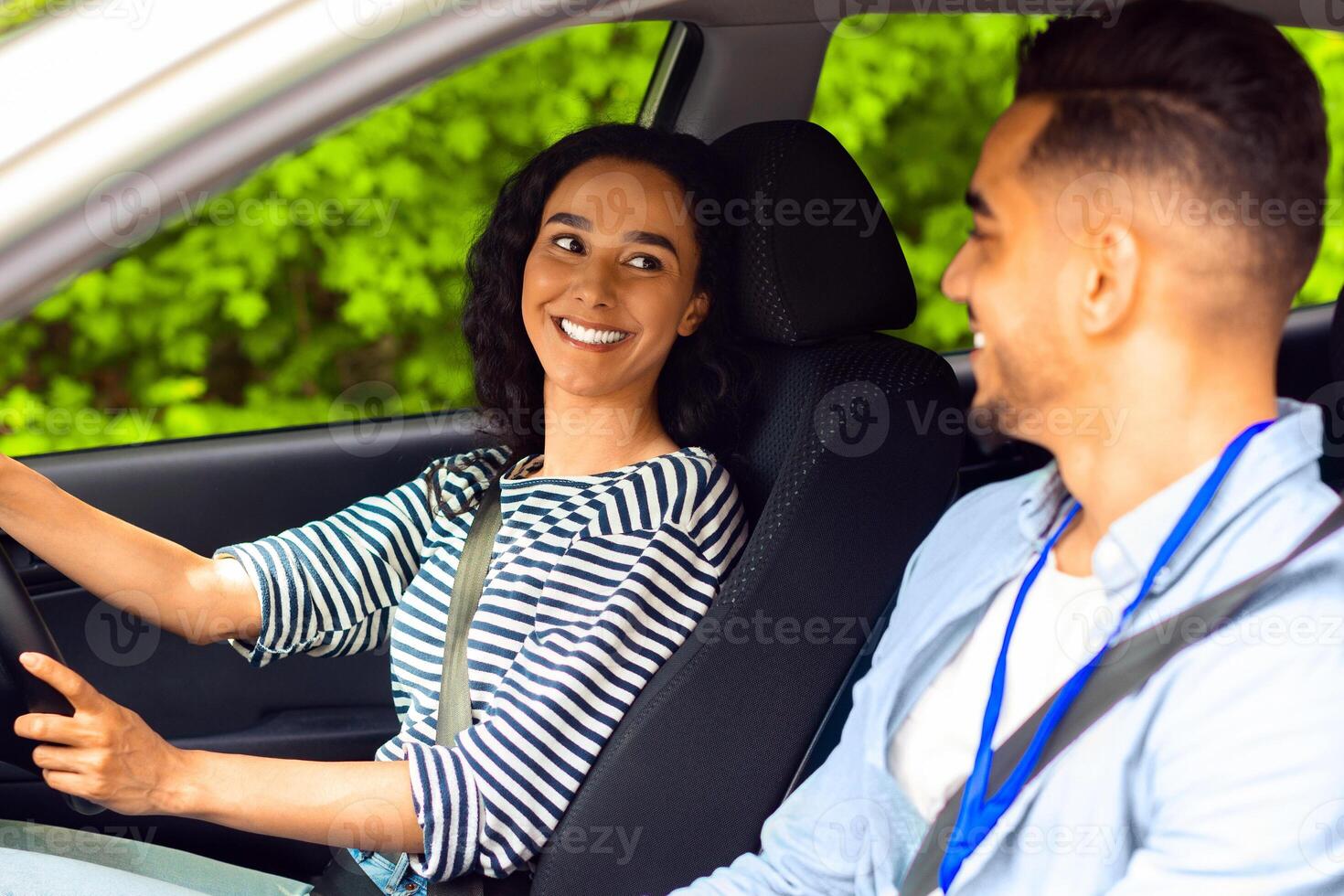 Excited young brunette woman sitting at driving car seat, looking at handsome arab man and smiling greeting instructor, female student of driving school ready for new lesson, side view photo