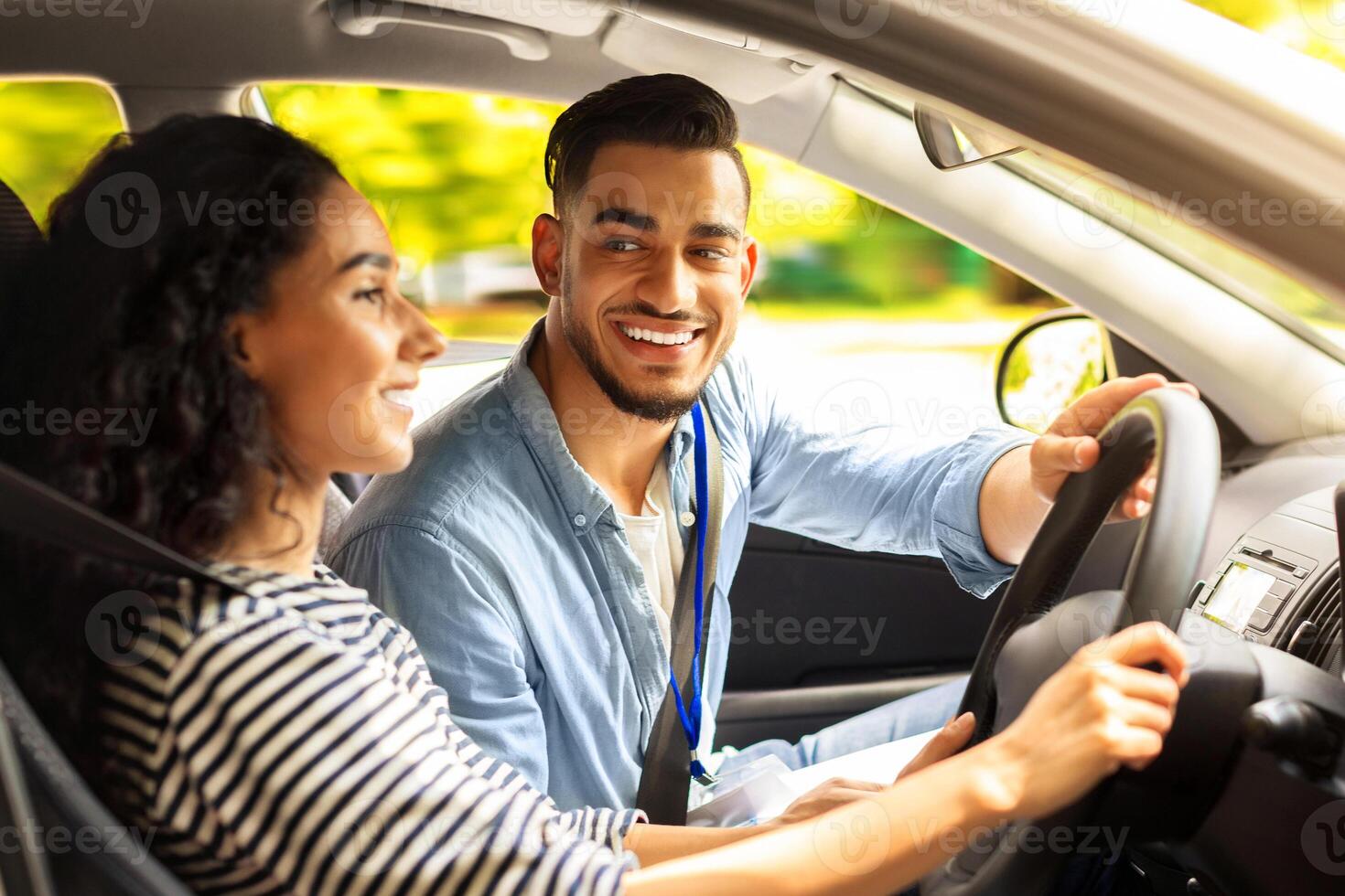 Handsome arab man instructor examinating brunette lady student, holding hand on steering wheel and looking at smiling young woman driving auto, side view, copy space. Driving school concept photo