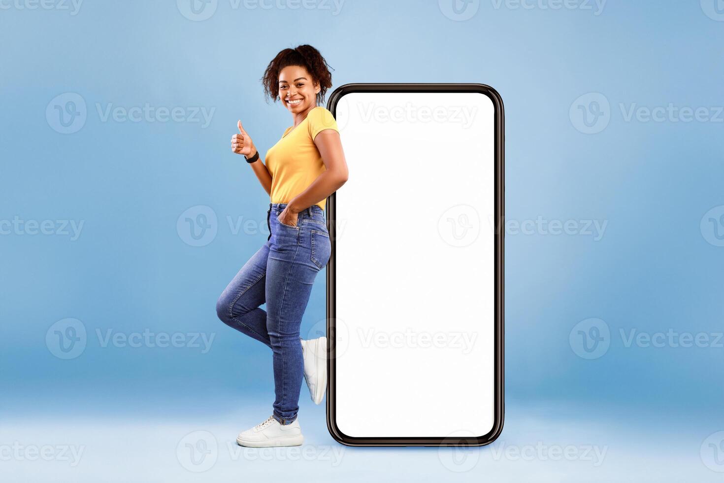 Pretty African American lady standing near big smartphone with blank screen, gesturing thumb up on blue background, mockup. Young woman recommending new mobile app, advertising website, free space photo