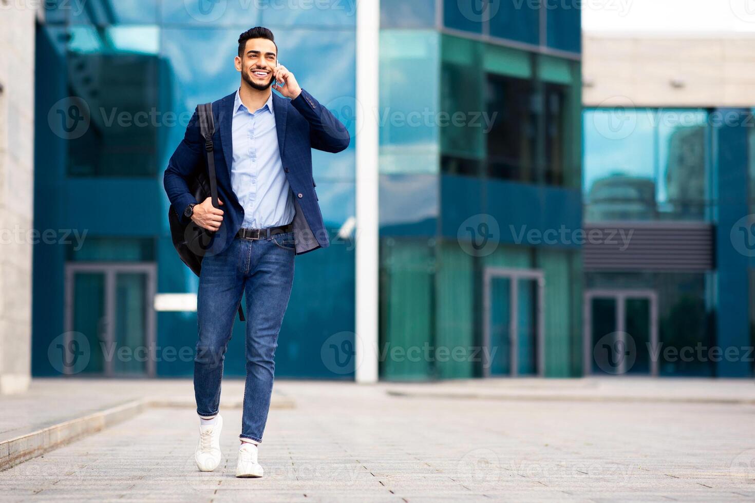 Young middle-eastern guy entrepreneur in formal clothes with backpack going home from office, talking on phone and smiling, having business conversation, business center background, copy space photo