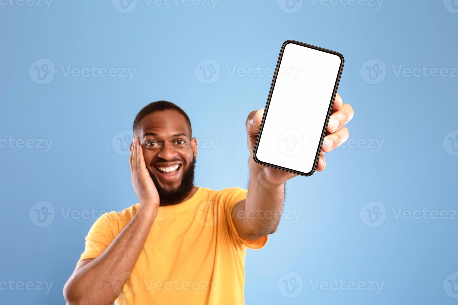 Check out this cool mobile app. Excited African American man presenting smartphone with empty screen, recommending website or ad on blue studio background, space for mockup. Selective focus photo