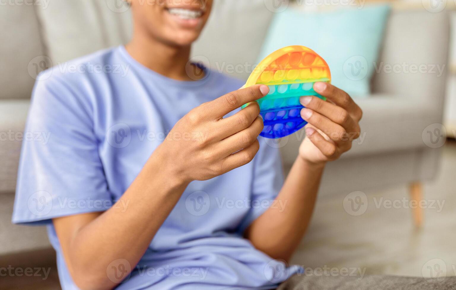 Unrecognizable black teen guy using POP IT toy to relieve stress at home, closeup. Cropped view of African American teenager playing with modern antistress fidget game, indoors photo