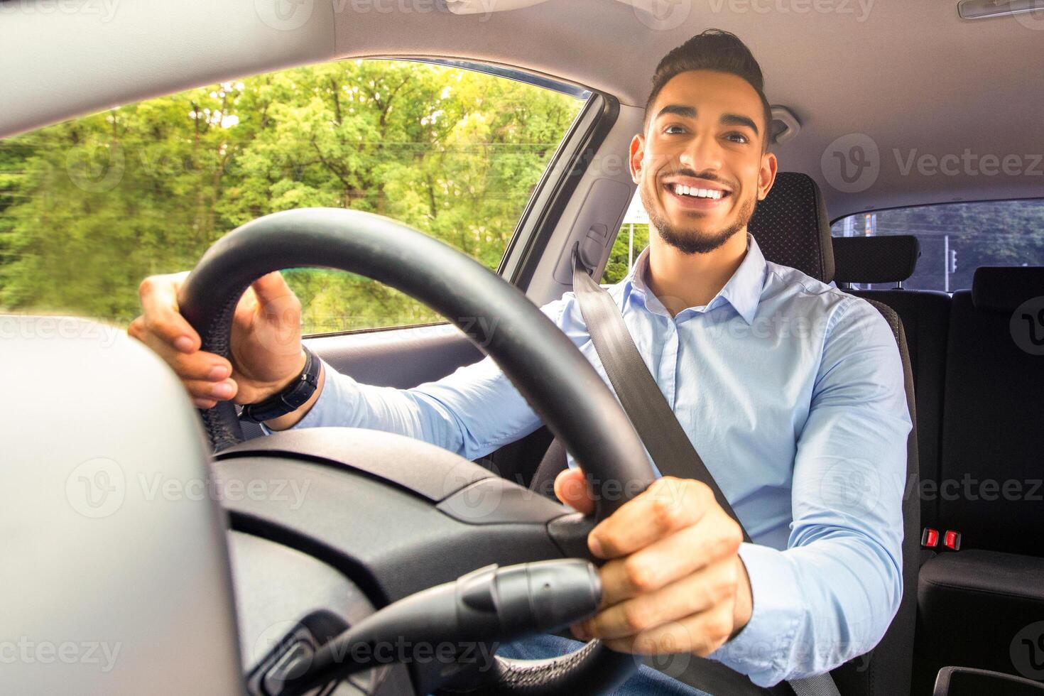 Smiling arab guy going to office in the morning, driving his car, copy space, shot from dashboard. Cheerful middle-eastern young man looking at the road, sitting inside luxury automobile, going home photo
