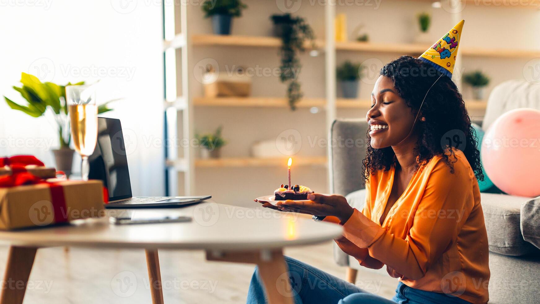 Online Birthday Celebration. Happy Black Woman In Festive Hat Blowing Candles On Cake Celebrating Having Distance Virtual Party With Friends Via Call On Laptop Computer At Home. Side View photo
