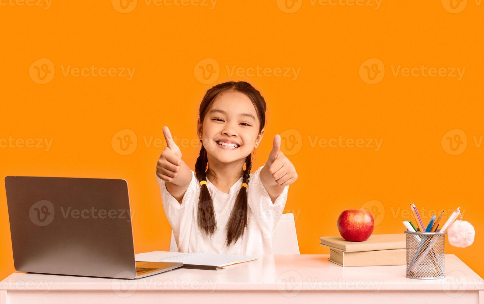 Smiling Asian Elementary School Girl At Laptop Gesturing Thumbs Up Sitting At Desk On Yellow Studio Background. I Like E-Learning. photo