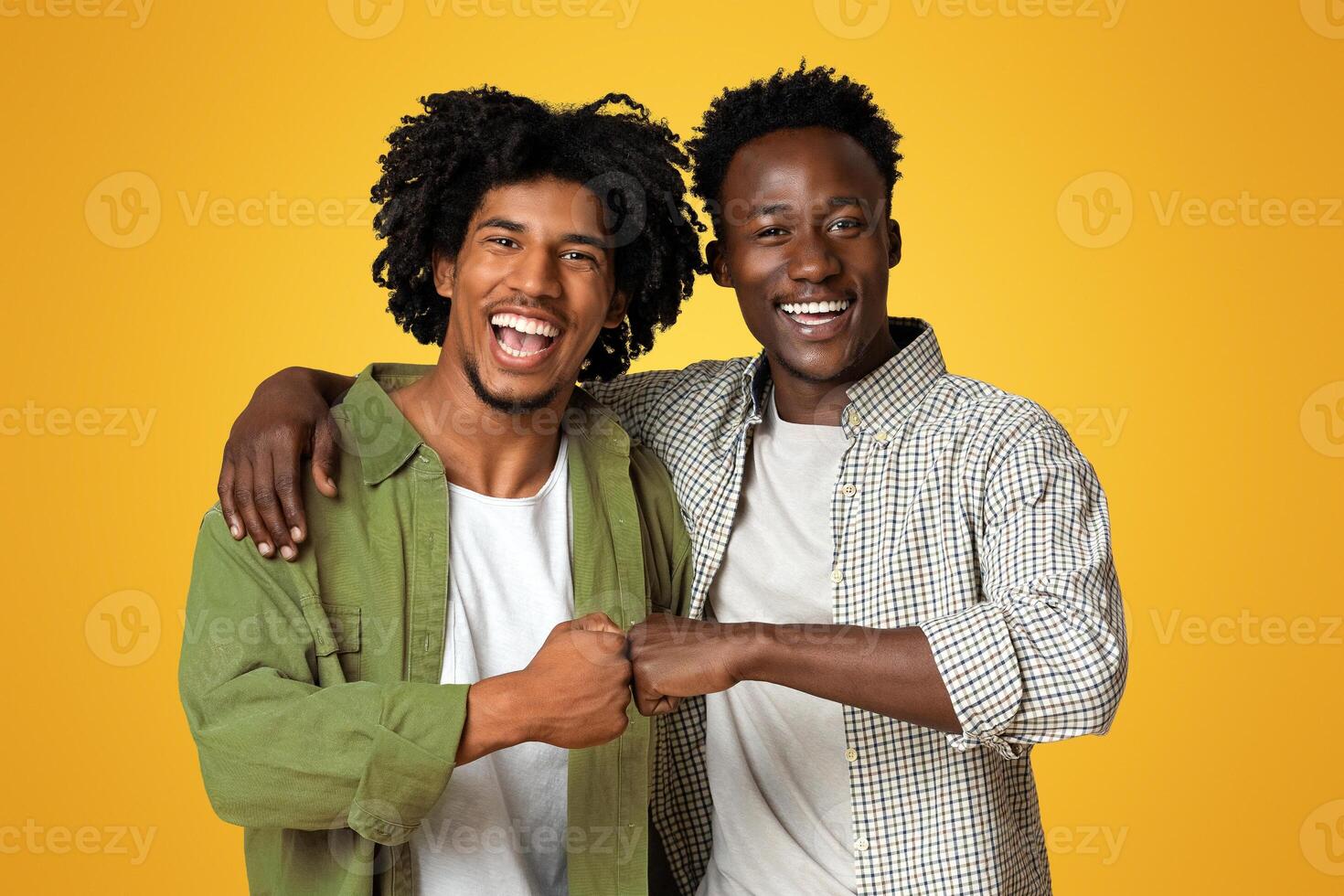 Portrait Of Two Cheerful Black Guys Embracing And Making Fist Bump Gesture, Happy Young African American Male Friends Having Fun Together While Standing Over Beige Studio Background, Copy Space photo