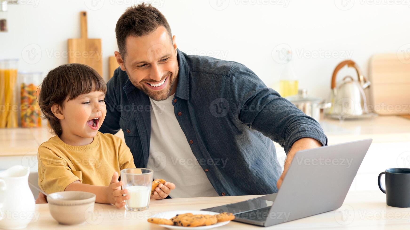 Happy young father with his little son sitting at kitchen and networking on laptop during breakfast, dad showing computer screen to interested kid, free space photo