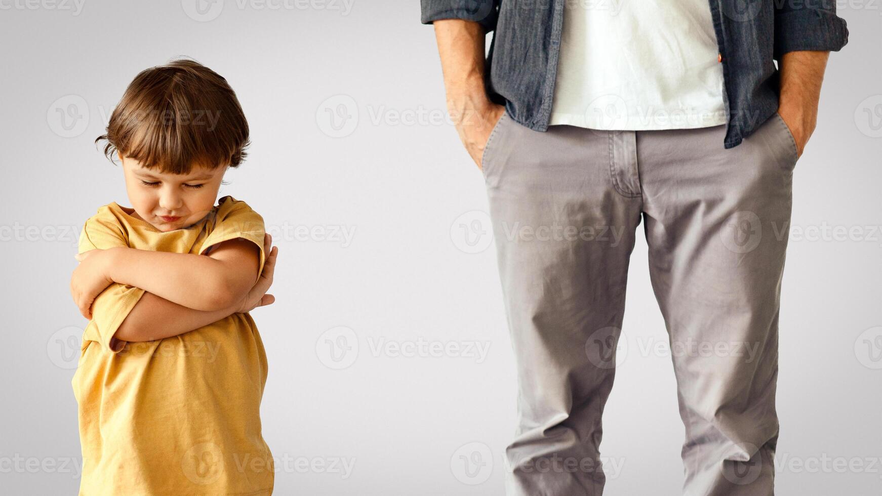 Kids caprices. Offended little boy standing with crossed hands, feeling resentment to his father, unrecognizable indifferent man standing behind with hands in pockets against white wall photo