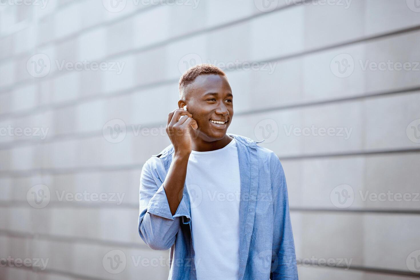 Cheerful black guy with earphones listening to his favorite music while walking near brick wall on city street photo