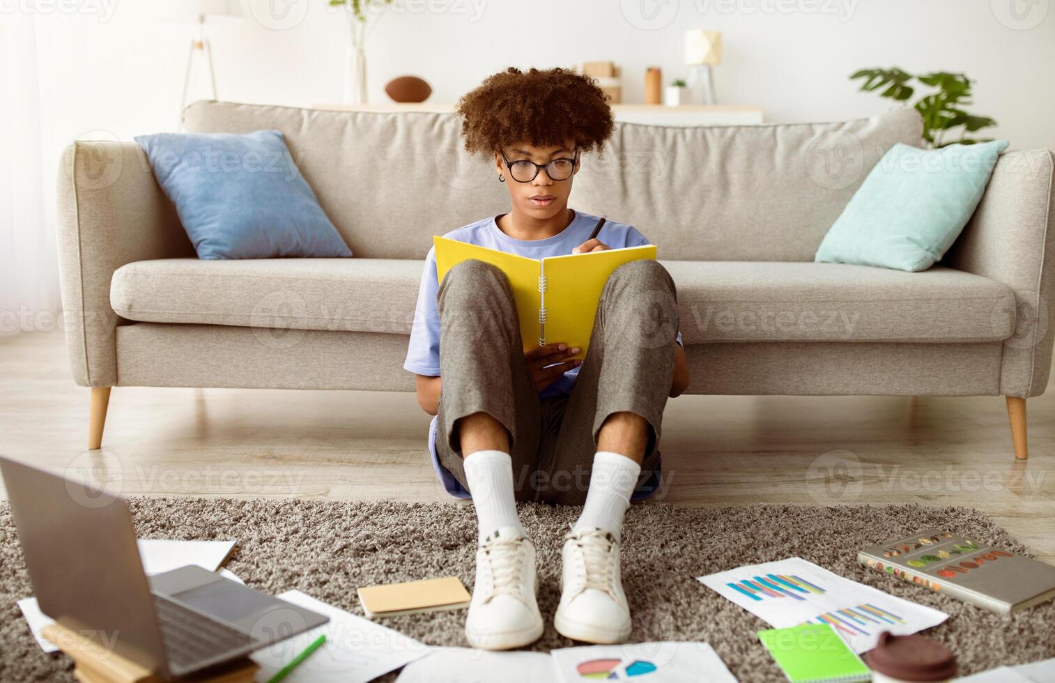 Web based education. Full length of black teenager writing down info during online class, sitting on floor in front of laptop at home. Afro teen guy having consultation with tutor, taking notes photo