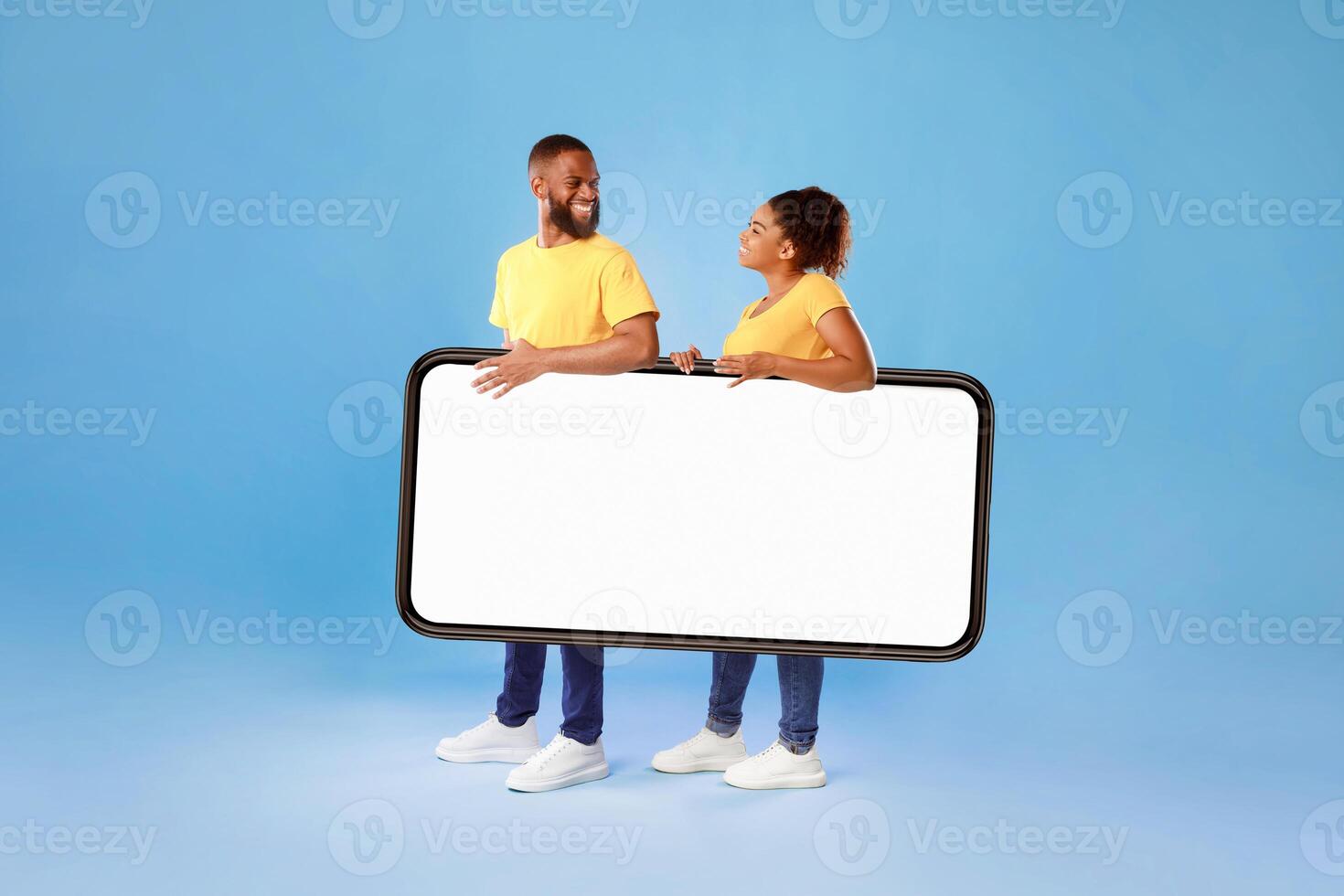 Positive African American couple carrying huge smartphone together, offering mockup for your app or website design on empty white screen, blue studio background. Cellphone ad template photo