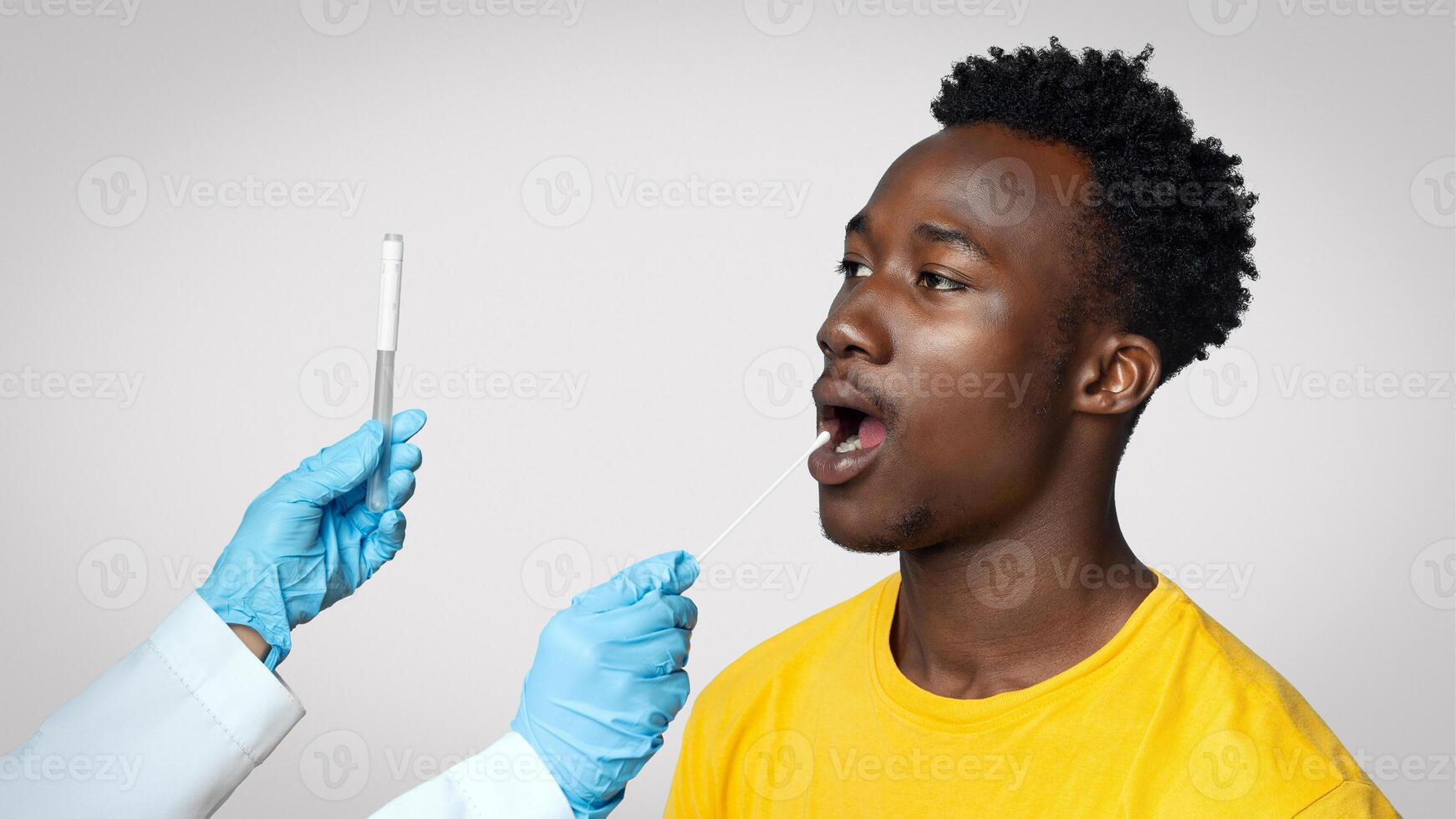 Doctor Holding Swab Making Oral PCR Coronavirus Test For Black Guy, Young African American Male Patient Undergoing Covid Diagnostic Procedure, Standing Over Blue Studio Background, Copy Space photo