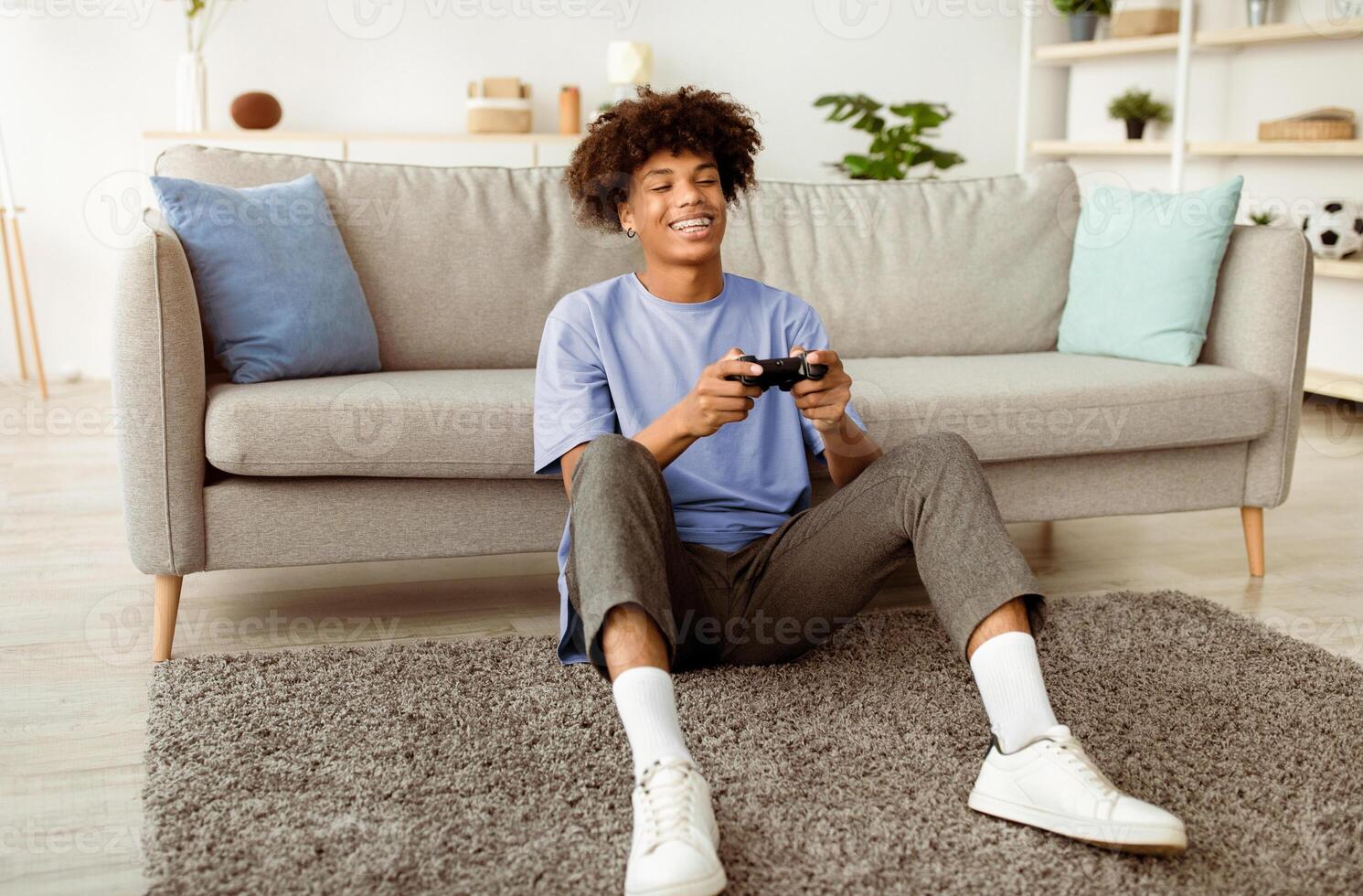 Happy black teenager enjoying videogame on playstation, having fun at home, full length. Joyful African American adolescent gamer with joystick sitting on floor, playing computer arcade photo