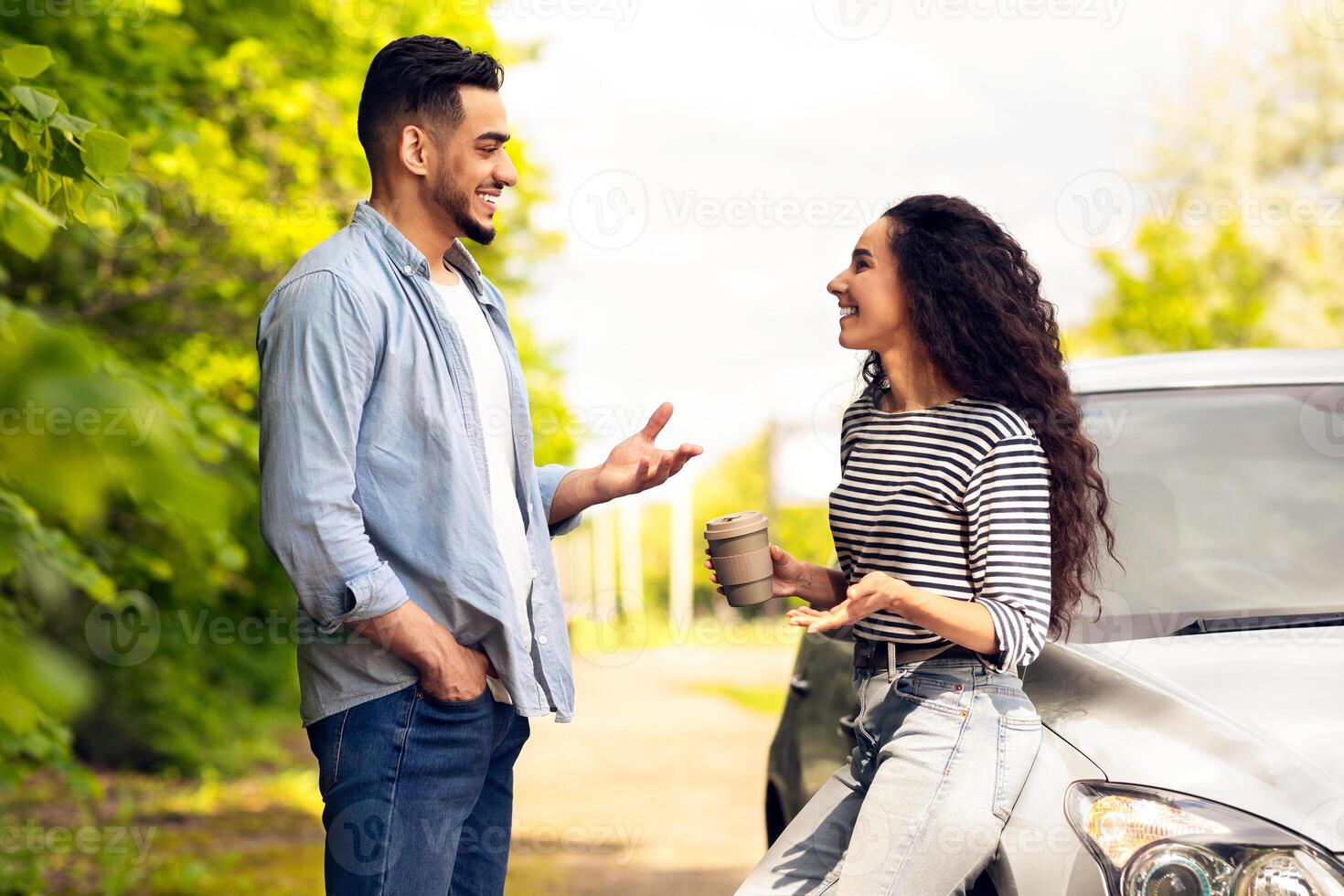 Happy couple having break while travelling by car together, beautiful young middle-eastern man and woman standing by auto at country side, drinking coffee and having conversation, side view photo