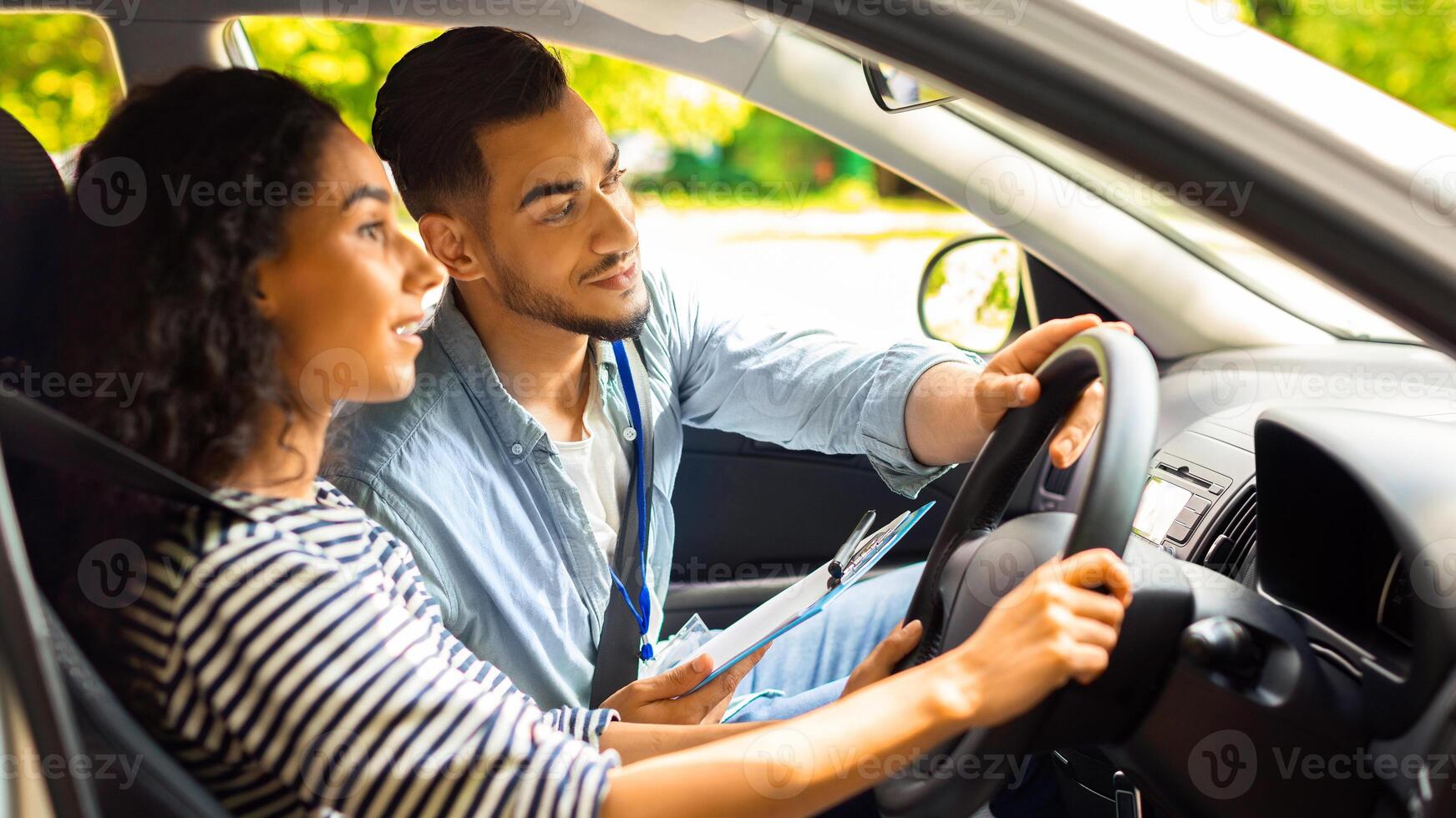 Handsome arab man driving instructor holding steering wheel, showing concentrated how to drive on the road, brunette lady young woman having lesson at driving school, side view, closeup photo