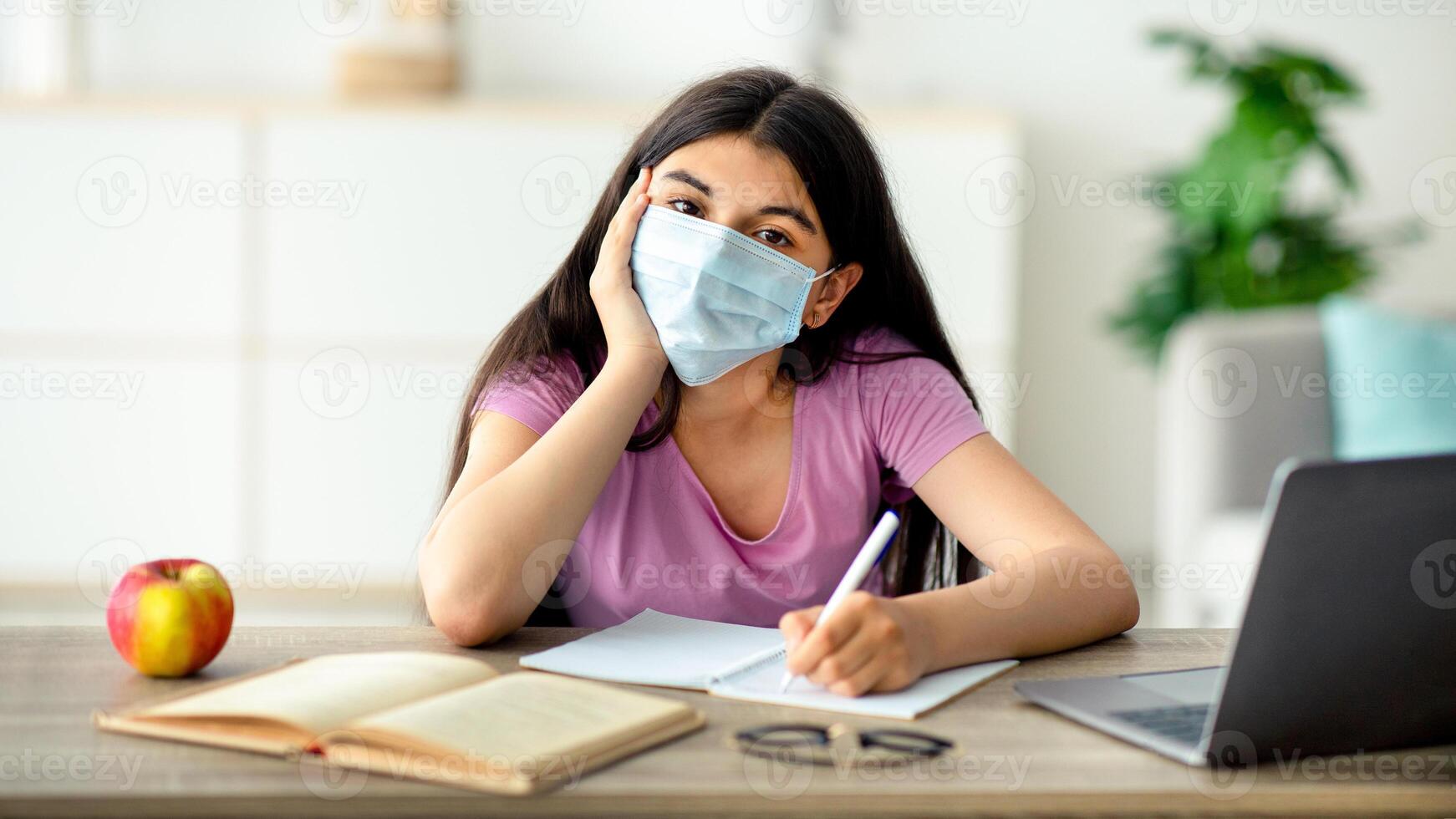 Remote education during coronavirus concept. Indian teen girl in face mask sitting at table with laptop, taking notes, making study project for online course, participating in web conference at home photo