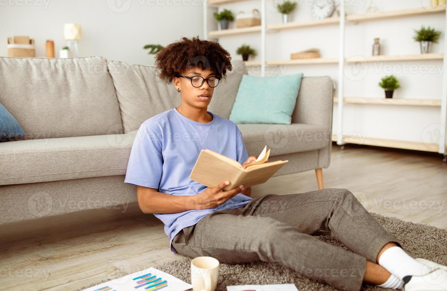 Focused black teenager reading book, sitting on floor, studying from home. Intelligent African American adolescent learning remotely, making assignment, using textbook to get ready for exam or test photo