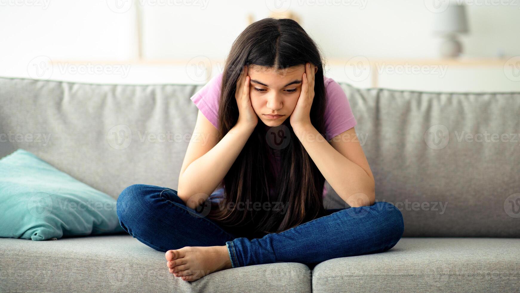 Covid related teen depression concept. Unhappy Indian teenage girl sitting on sofa at home with head in her hands, suffering from lack of communication and loneliness, feeling stressed during lockdown photo